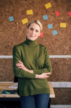 Portrait of a female office worker. She is standing in front of a cork board, smartly dressed with her arms folded and is smiling for the camera.