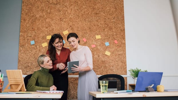 Women are working together in an office. One of the women is holding a digital tablet which they are all looking at.