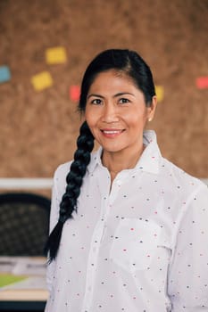 Portrait of a beautiful woman in her workplace. She is standing in front of a corkboard and is smartly dressed, smiling for the camera.
