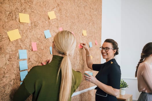 Two women are at work in an office. They are standing at a cork board and are having a discussion as they pin things up.