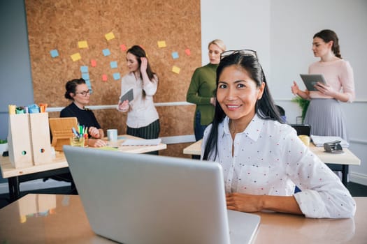 Beautiful woman is smiling for the camera while sitting at her desk in her workplace. There are women working in the background.