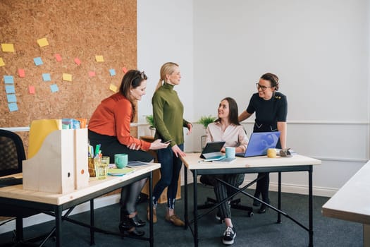 Women are working together in an office. They are talking as they use technology.