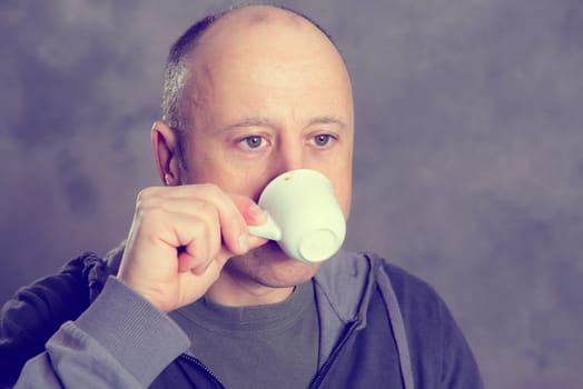 young baldheaded man drinking coffee in front of gray background