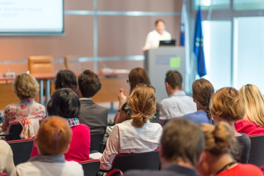 Business and entrepreneurship symposium. Speaker giving a talk at business meeting. Audience in conference hall. Rear view of unrecognized participant in audience.