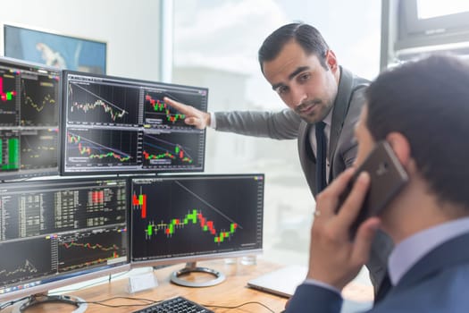 Businessmen trading stocks online. Stock brokers looking at graphs, indexes and numbers on multiple computer screens. Colleagues in discussion in traders office. Business success concept.