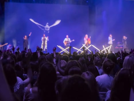 Audience with hands raised at a music festival and lights streaming down from above the stage. Focus on hands.