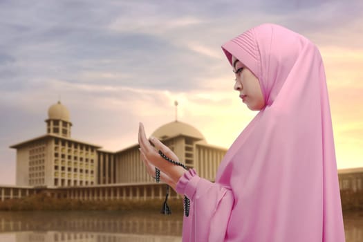 Young asian muslim woman praying with prayer beads against mosque background