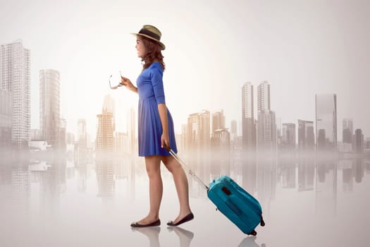Young asian woman with hat holding suitcase over cityscape background