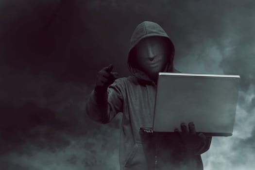 Portrait of hooded hacker with mask holding laptop while standing against dark room