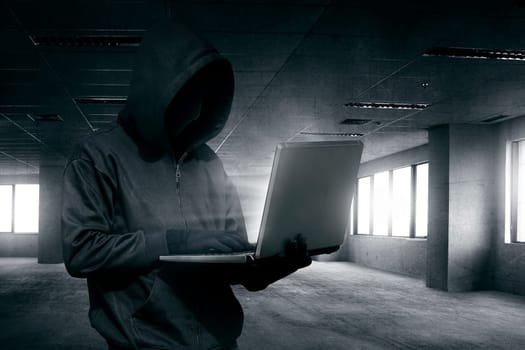 Hooded man with mask using laptop to hacking on the empty dark room