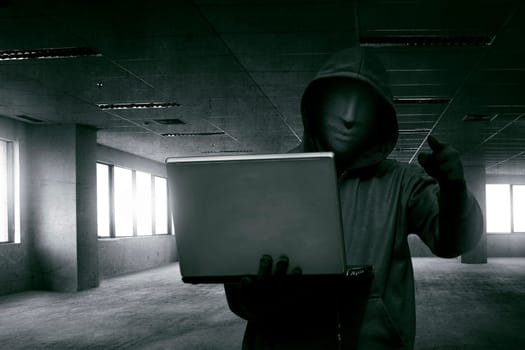 Hacker man with anonymous mask holding laptop while standing against dark room