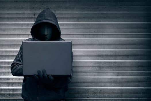 Hooded hacker with mask holding laptop while typing against metal roller shutter door background