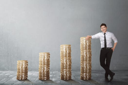 Confident asian businessman standing beside coin stack against grey wall background