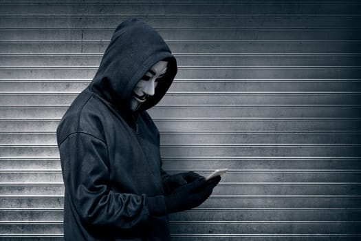 Hooded man with vendetta mask using mobile phone against roller shutter background