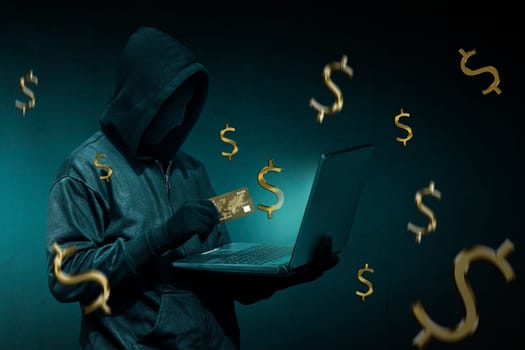 Hooded hacker with credit card stealing data from a laptop over dark background