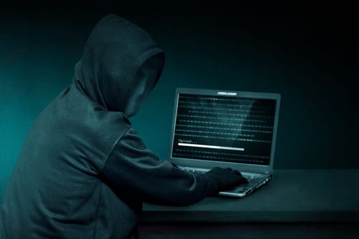 Hooded hacker with anonymous mask using laptop to steal data in the dark room
