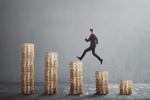 Happy asian businessman jump on between coin stacks against grey background