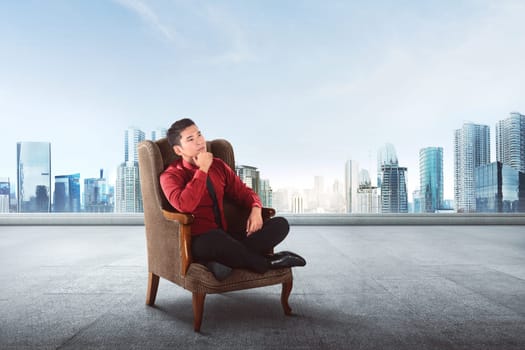 Thoughtful asian businessman sitting on sofa thinking about something with skyscraper background