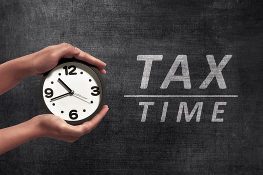 People hand holding black alarm clock beside Tax time announcement on blackboard