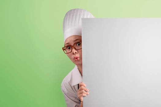Funny asian woman chef peeking from behind blank copyspace over green background