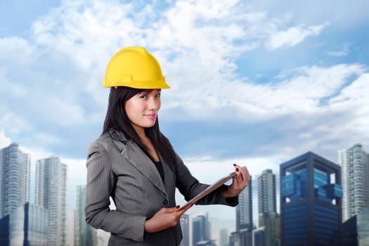 Smiling asian contractor woman with yellow helmet holding clipboard against skyscraper background