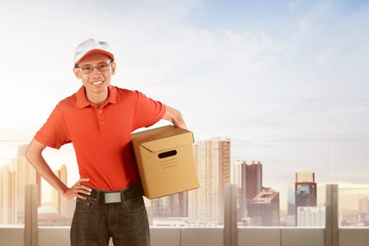 Happy asian courier holding big parcel against cityscape background