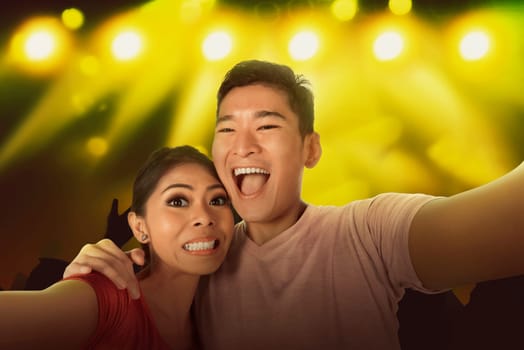 Young asian couple selfie with the crowd in front of bright stage lights
