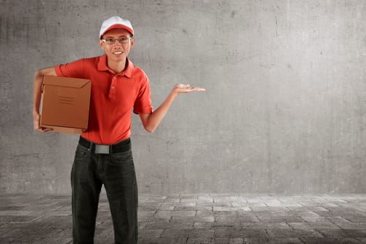 Smiling asian courier man delivery holding package with open arm against gray wall background