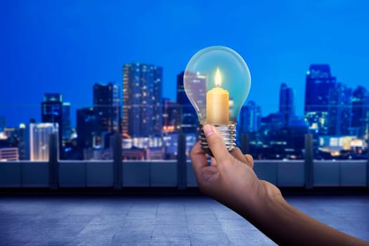 Hand holding light bulb with shining candle inside it on modern city background