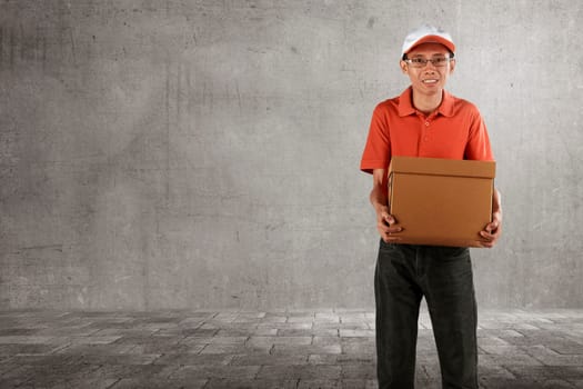 Cheerful asian courier standing with parcel over grey wall background