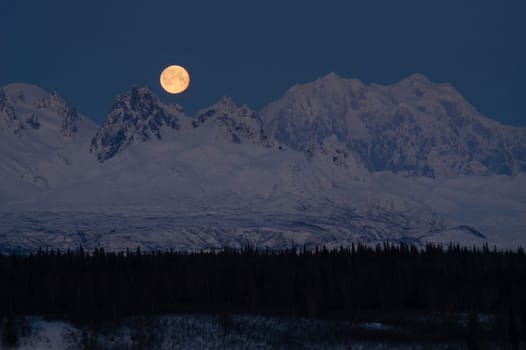 The moon emerges from behind the mountains in Denali