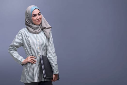 Happy asian muslim woman wearing veil holding laptop against dark background