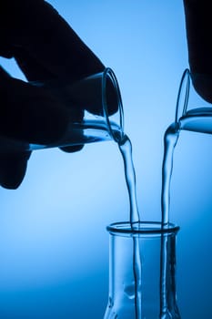 Two hand sihouettes,each with a test tube,fill an Erlmeyer flask against a blue-tinted studio background.
