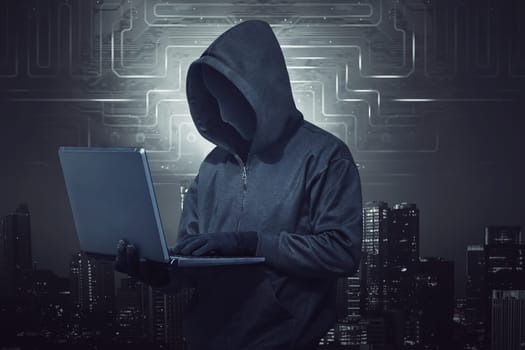 Hooded hacker holding laptop while typing against digital city background