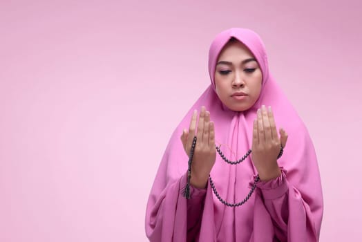 Young asian muslim woman praying with prayer beads over pink background