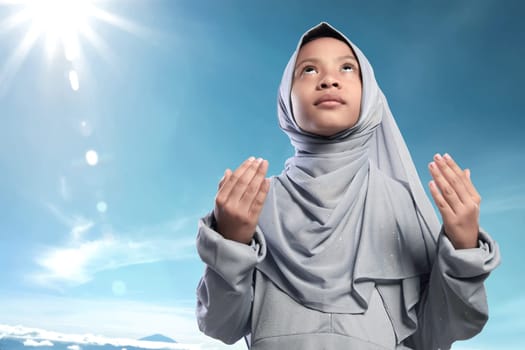 Religious asian muslim kid raising hand and praying against blue sky background