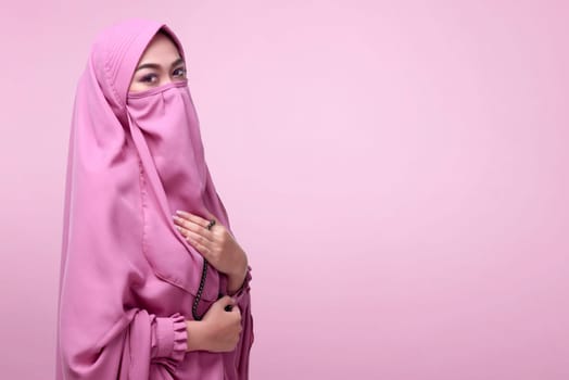 Young asian muslim woman in veil with prayer beads standing over pink background