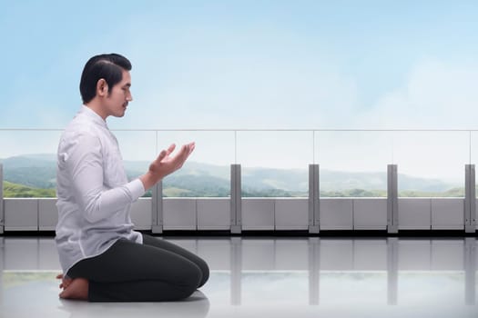 Young asian muslim man sitting in the terrace praying with blue sky background