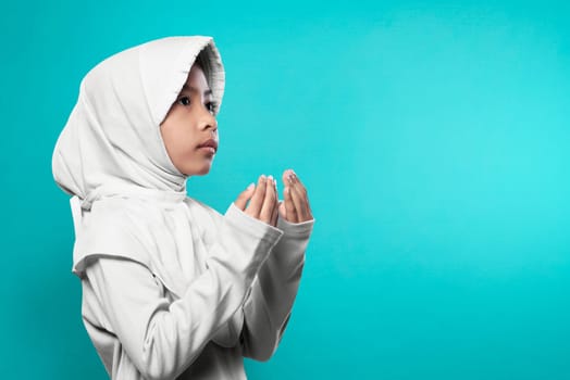 Asian muslim girl in hijab raising hand and praying over blue background