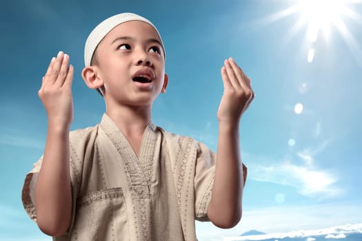 Cheerful asian muslim child in traditional dress praying with blue sky background