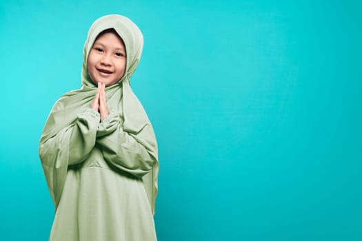 Smiling asian muslim child with hijab dress praying to over blue background
