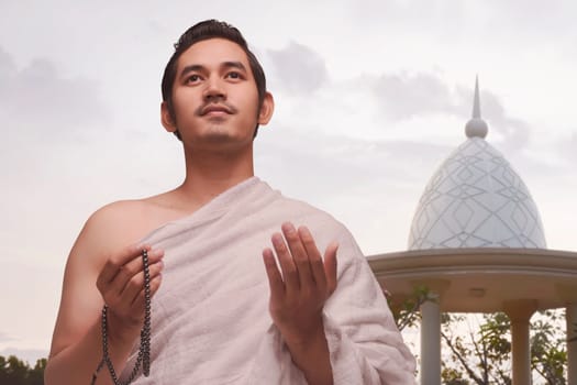 Smiling asian muslim man with ihram clothes holding prayer beads over sky background