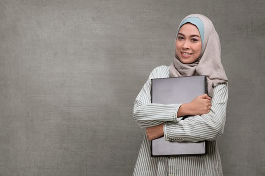 Beautiful asian muslim woman holding laptop against concrete background