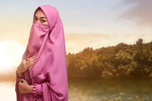 Asian muslim woman in veil with prayer beads standing with sunset background