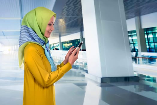 Pretty asian muslim woman touching her mobile phone against indoor building background