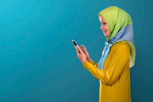 Beautiful asian muslim woman concentrates while using her smartphone on blue background