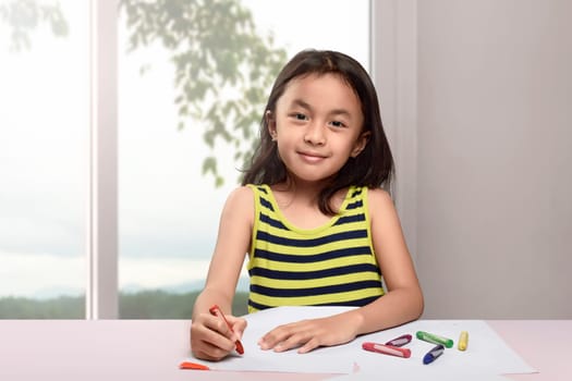 Happy asian child drawing with crayon on white paper in the home