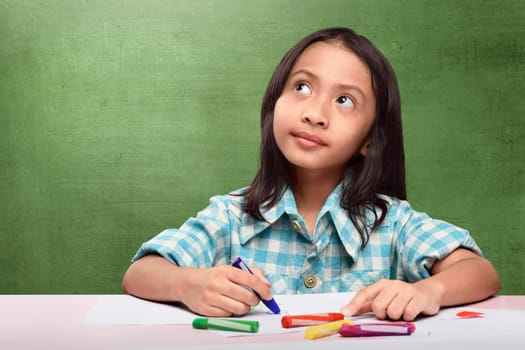Cheerful asian child imagining something to drawing with crayon at school