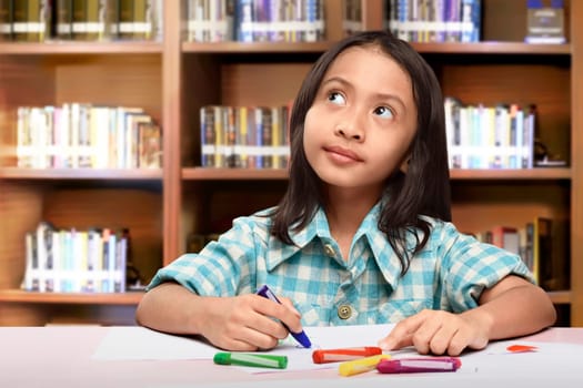 Cheerful asian kid girl with crayon want to drawing something against bookshelf background