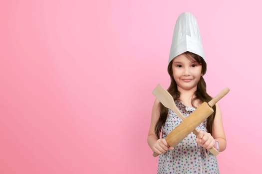 Smiling asian little girl in chef hat holding wooden cooking utensils over pink background